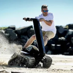 Mężczyzna w okularach słonecznych na pojeździe gąsienicowym, piasek w ruchu, skały w tle, jasne niebo.