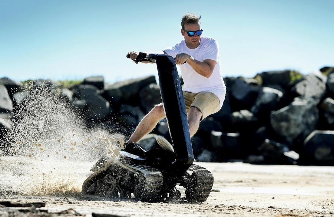 Mężczyzna w okularach słonecznych na pojeździe gąsienicowym, piasek w ruchu, skały w tle, jasne niebo.