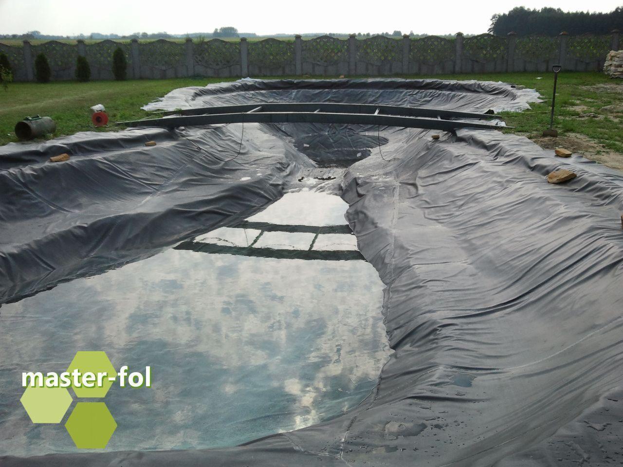 Wykładanie folią dna oczka wodnego w trakcie budowy, widoczne drewniane wzmocnienia, częściowo wypełnione wodą, z logo producenta folii w rogu kadru.
