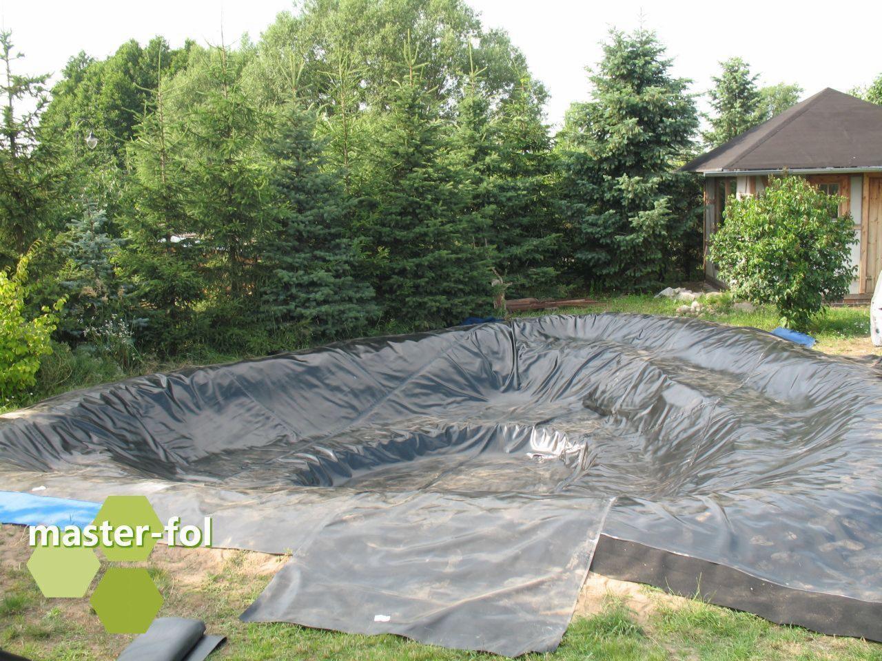 Wykładanie folii uszczelniającej w przygotowanym wykopie pod oczko wodne na tle zielonych drzew iglastych i drewnianego domku.