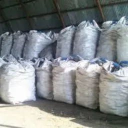 Rzędy worków z pelletem pod zadaszeniem magazynu. Worki ułożone jeden na drugim, widoczne szwy. Oświetlenie naturalne, blaszany dach.