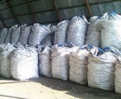 Rzędy worków z pelletem pod zadaszeniem magazynu. Worki ułożone jeden na drugim, widoczne szwy. Oświetlenie naturalne, blaszany dach.