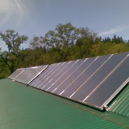 Rząd paneli słonecznych zamontowanych na zielonym, blaszanym dachu z widokiem na zielone drzewa i błękitne niebo z chmurami.