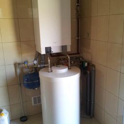 TWORKOWSKI S.C. - Biały, wiszący piec gazowy Viessmann z podłączonym zasobnikiem ciepłej wody w kotłowni wyłożonej beżowymi płytkami ceramicznymi. Widoczne rury instalacji grzewczej, naczynie przeponowe i kartonowe...