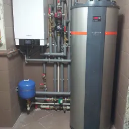 W kotłowni zainstalowany biały piec gazowy Viessmann obok szarego zbiornika na wodę Viessmann Vitocal 160 z pomarańczowymi akcentami, połączone szarymi rurami z zaworami.