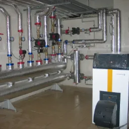 Instalacja kotłowni z widocznym kotłem gazowym De Dietrich, orurowaniem z izolacją termiczną, zaworami i manometrami na tle wykafelkowanej ściany i podłogi.