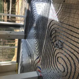 Instalacja ogrzewania podłogowego z rurami ułożonymi w spiralny wzór na folii z siatką, widoczne okno i elementy konstrukcyjne budynku.