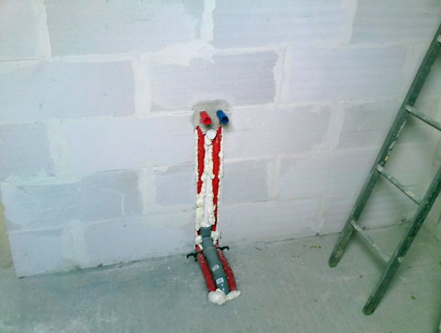 Wystające ze ściany z betonu komórkowego rury instalacji wodnej, czerwone i szare, zabezpieczone pianką montażową, widoczne niebieskie i czerwone końcówki, obok drabina.