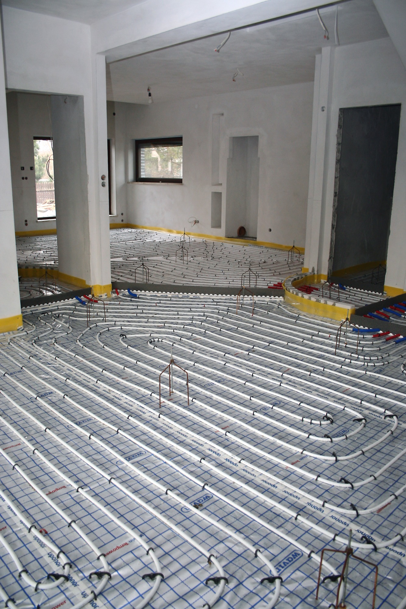 Rozległa instalacja ogrzewania podłogowego w surowym stanie, rury grzewcze ułożone na folii z siatką, widoczne mocowania i żółta taśma izolacyjna wzdłuż ścian.