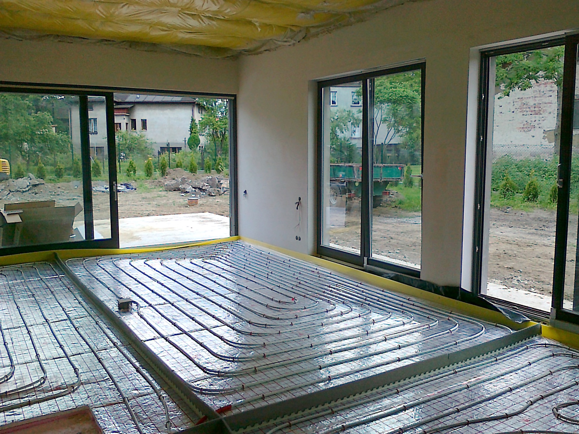 Instalacja ogrzewania podłogowego w nowym domu z dużymi oknami, rury grzewcze ułożone na folii aluminiowej z siatką, widoczne elementy mocujące i żółta taśma brzegowa.