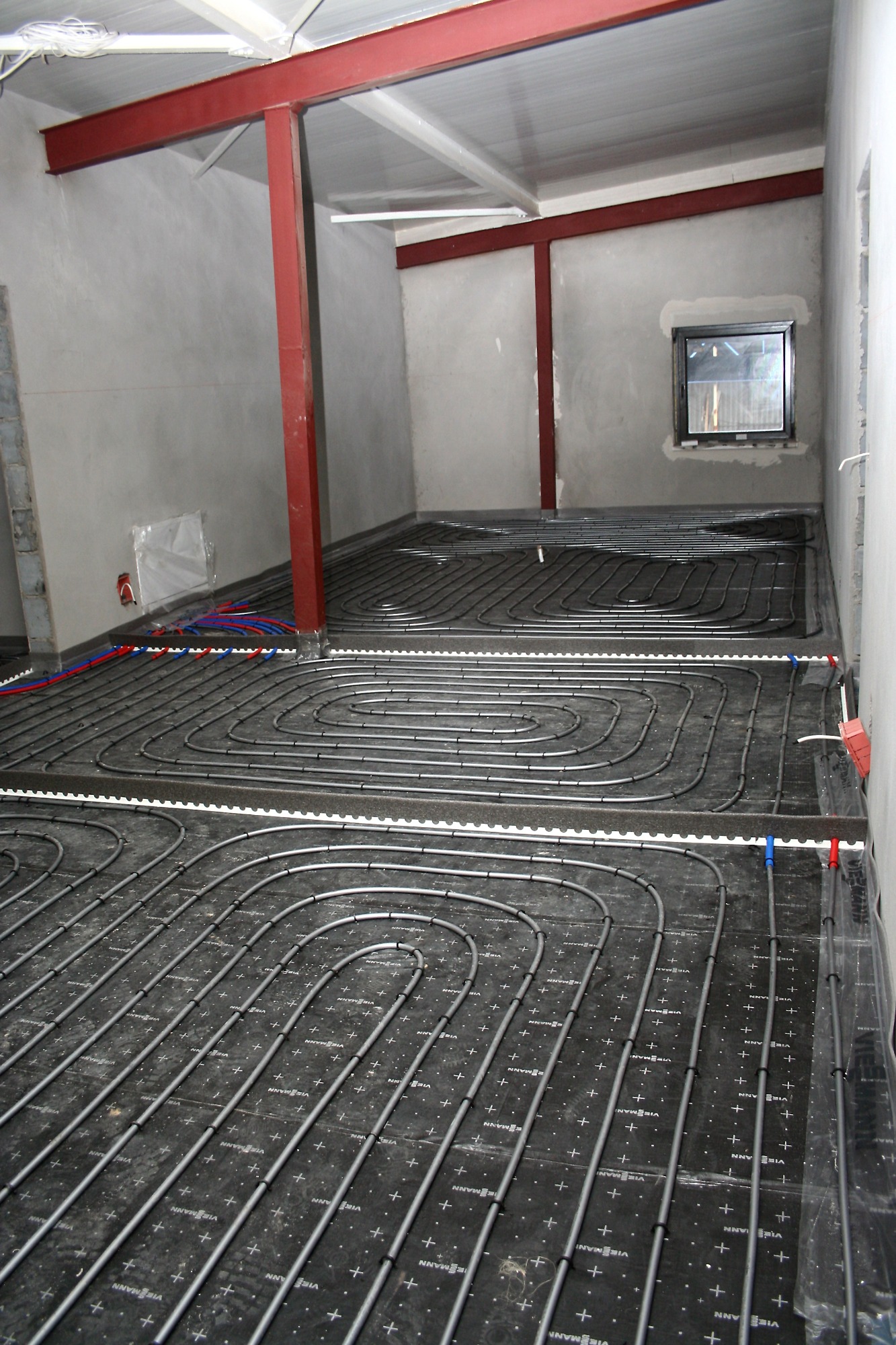 Wnętrze w trakcie montażu ogrzewania podłogowego: rury grzewcze ułożone spiralnie na czarnej macie z oznaczeniami, widoczne czerwone stalowe słupy konstrukcyjne, surowe ściany w szarym odcieniu...
