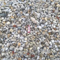 Kupa drobnego, jasnego kruszywa w Mińsku Mazowieckim, z pojedynczym czerwonym zapalniczką dla skali w centralnej części kadru, widoczne kamienie o różnych odcieniach szarości, beżu i brązu.