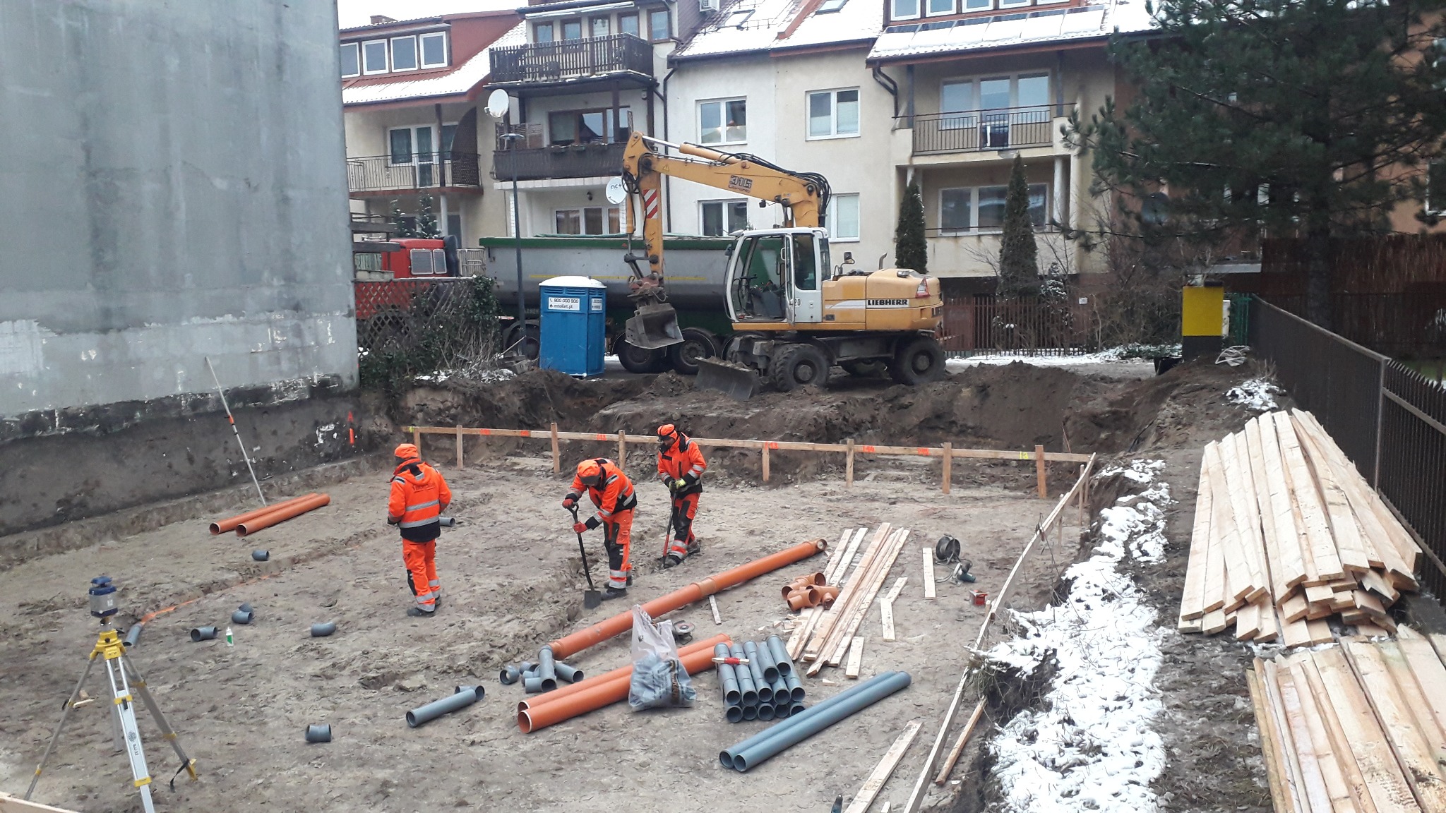 Przygotowanie terenu pod budowę: robotnicy w pomarańczowych kombinezonach kopią fundamenty, w tle koparka Liebherr i przenośna toaleta, widoczne rury kanalizacyjne i drewniane szalunki.