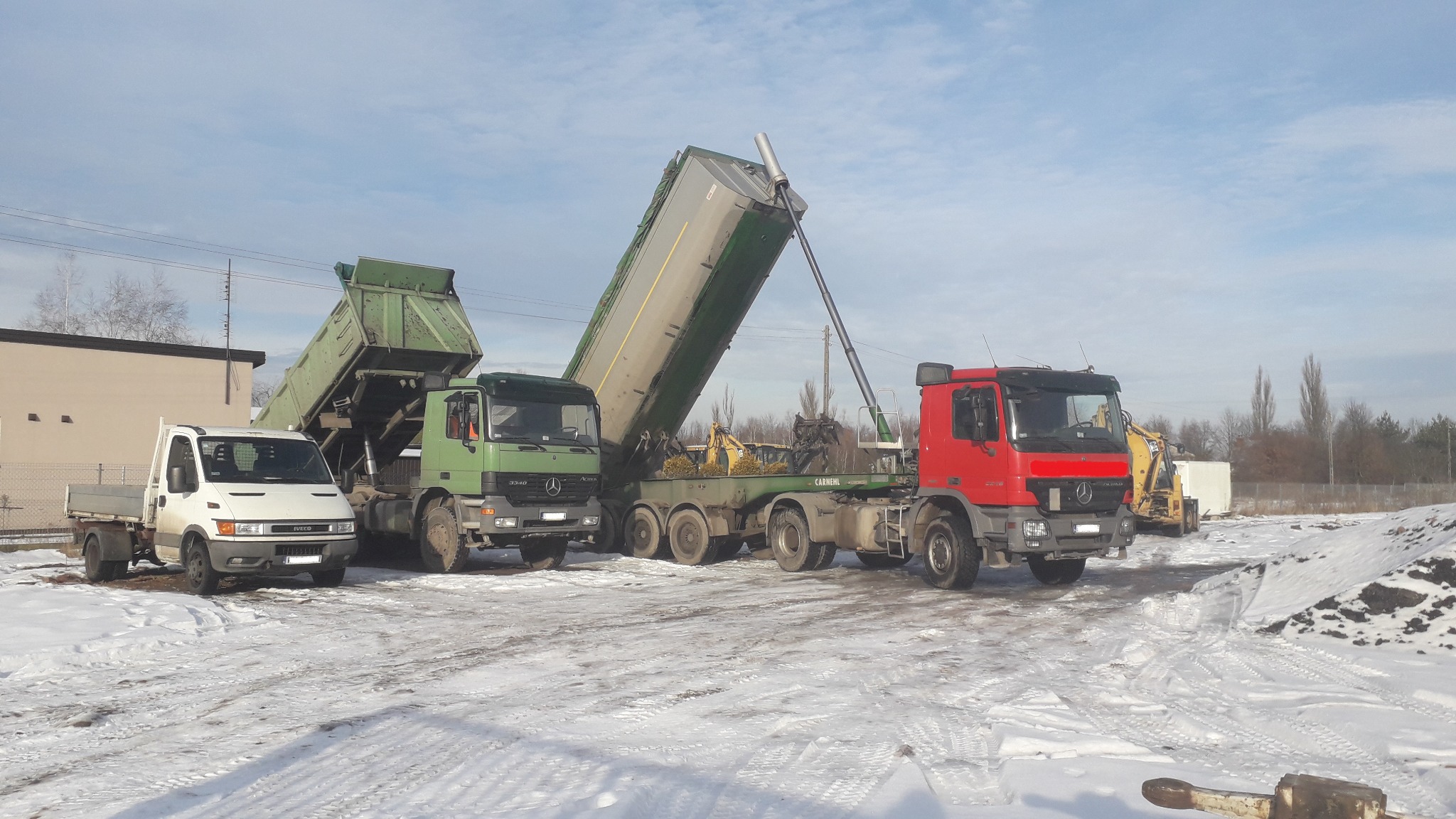 Rząd wywrotek: biała Iveco, zielony Mercedes z podniesioną skrzynią, zielony Mercedes i czerwony Mercedes, wszystkie stoją na śniegu, w tle budynek i drzewa.