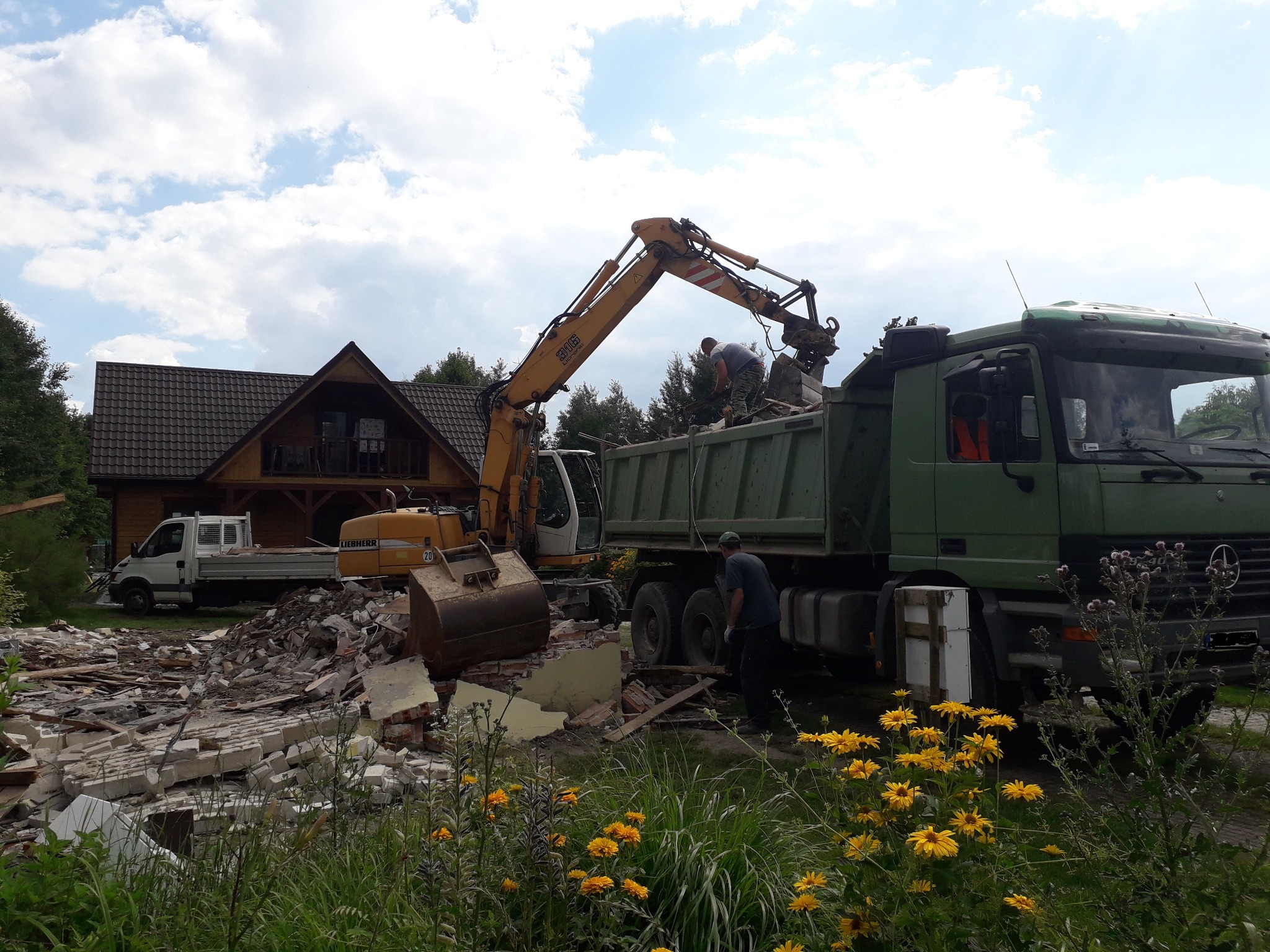 Żółta koparka Liebherr załadowuje gruz z rozbiórki ceglanego budynku do zielonej ciężarówki Mercedes-Benz; w tle drewniany dom z brązowym dachem i biały samochód dostawczy.