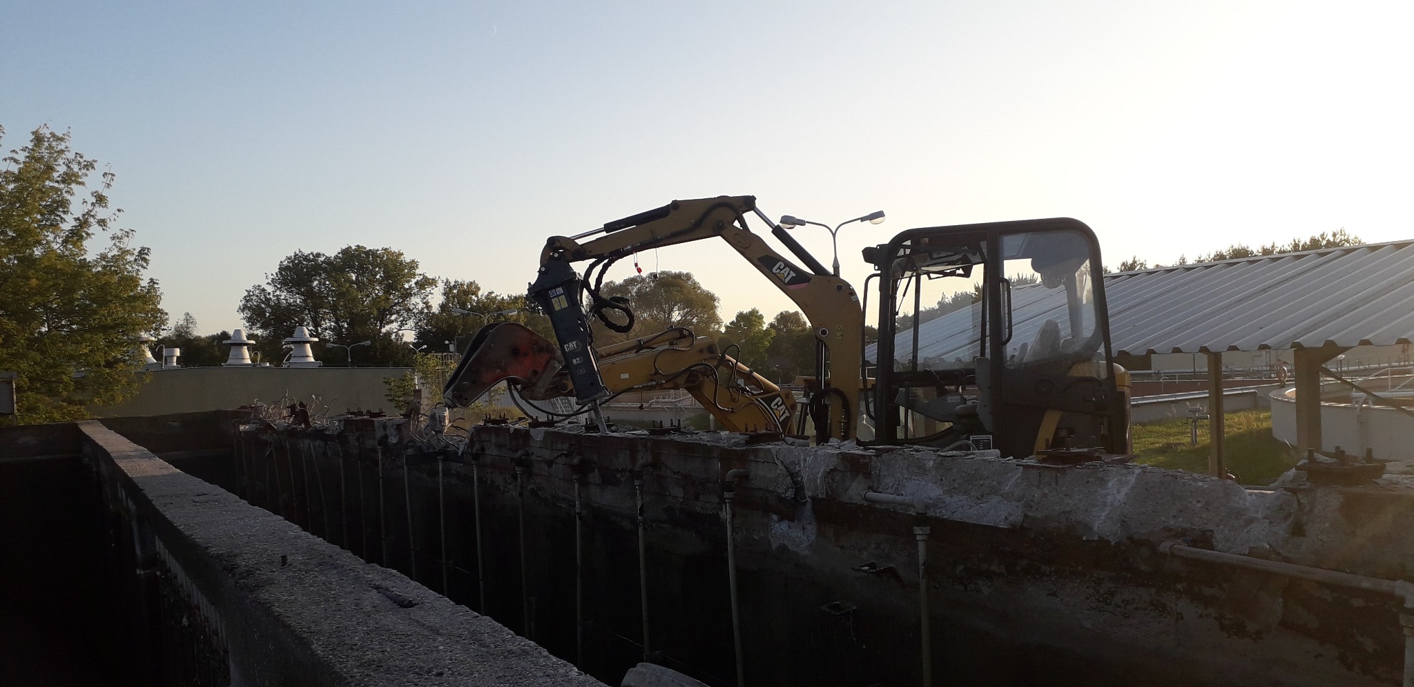 Żółta koparka CAT z młotem hydraulicznym kruszy betonową konstrukcję, prawdopodobnie zbiornik, w tle budynek przemysłowy i drzewa, wczesny ranek.