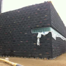 Czarny budynek z cegły klinkierowej z kolorowymi akcentami, widok z dołu. Część budynku jest osłonięta folią. Na dole widoczny żółty wąż i palety z cegłami.