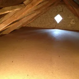 Podłoga na poddaszu z oknem w kształcie rombu, drewniana konstrukcja dachu. Widok na jasną, gładką powierzchnię podłogi i ceglaną ścianę.