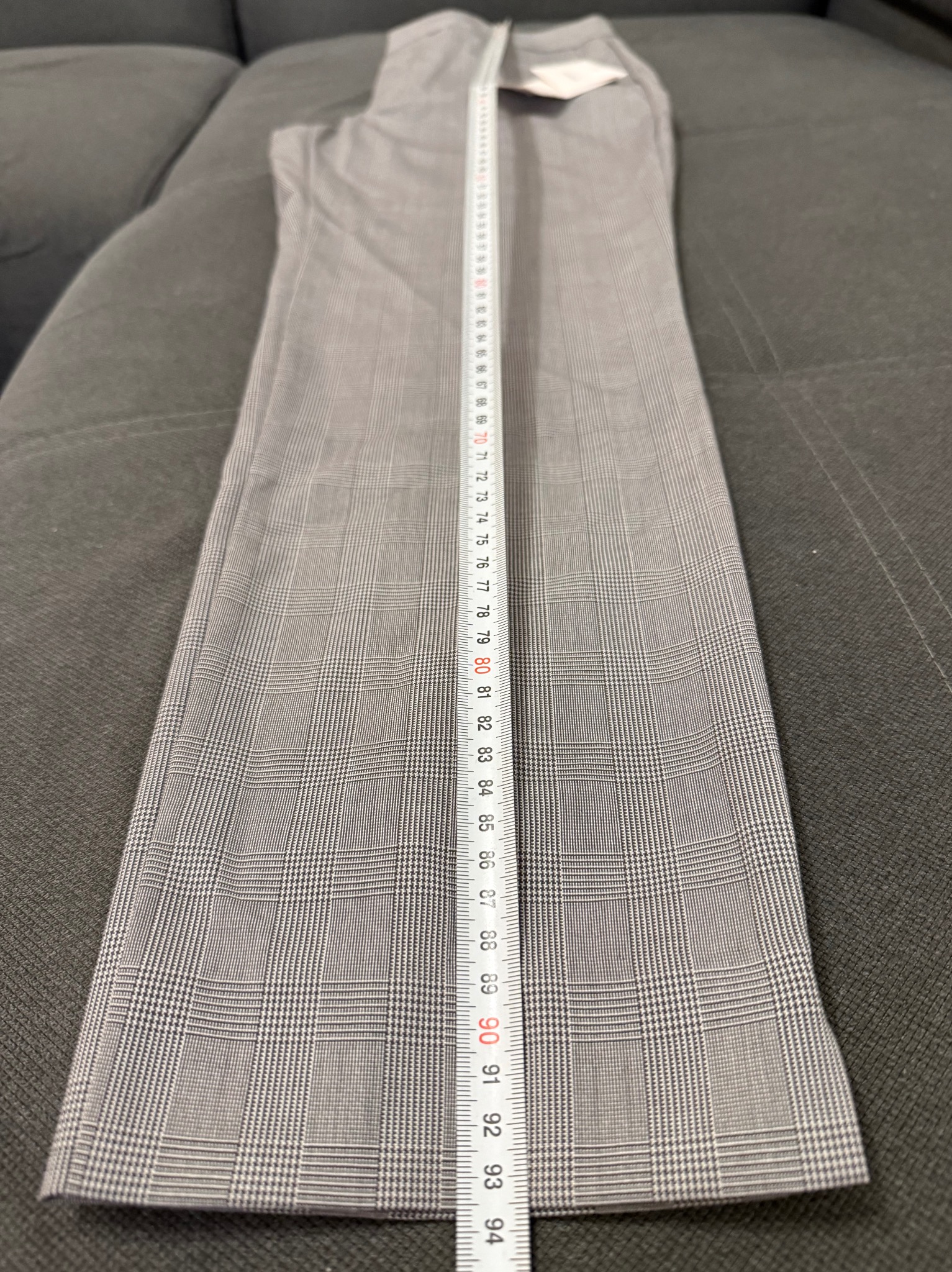 Szare spodnie w kratę leżące na kanapie, mierzone białą miarką. Widoczny wzór kraty i numeracja na miarce.