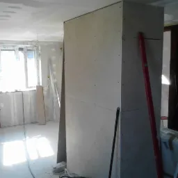 Wnętrze w trakcie prac z płyt karton-gips, widoczna ściana działowa i fragment okna. Czerwona poziomica oparta o ścianę. Częściowo widoczna drabina i osoba w tle.