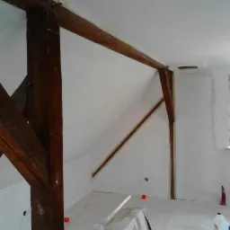 Białe ściany poddasza z drewnianymi belkami, po malowaniu. Okno PCV, zabezpieczona podłoga, widoczne ślady po taśmie malarskiej. Oświetlenie naturalne i sztuczne.