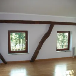 Jasne wnętrze poddasza z drewnianą podłogą, dwoma oknami z widokiem na drzewa i dekoracyjną, ciemną belką stropową. Grzejnik pod oknem po prawej.