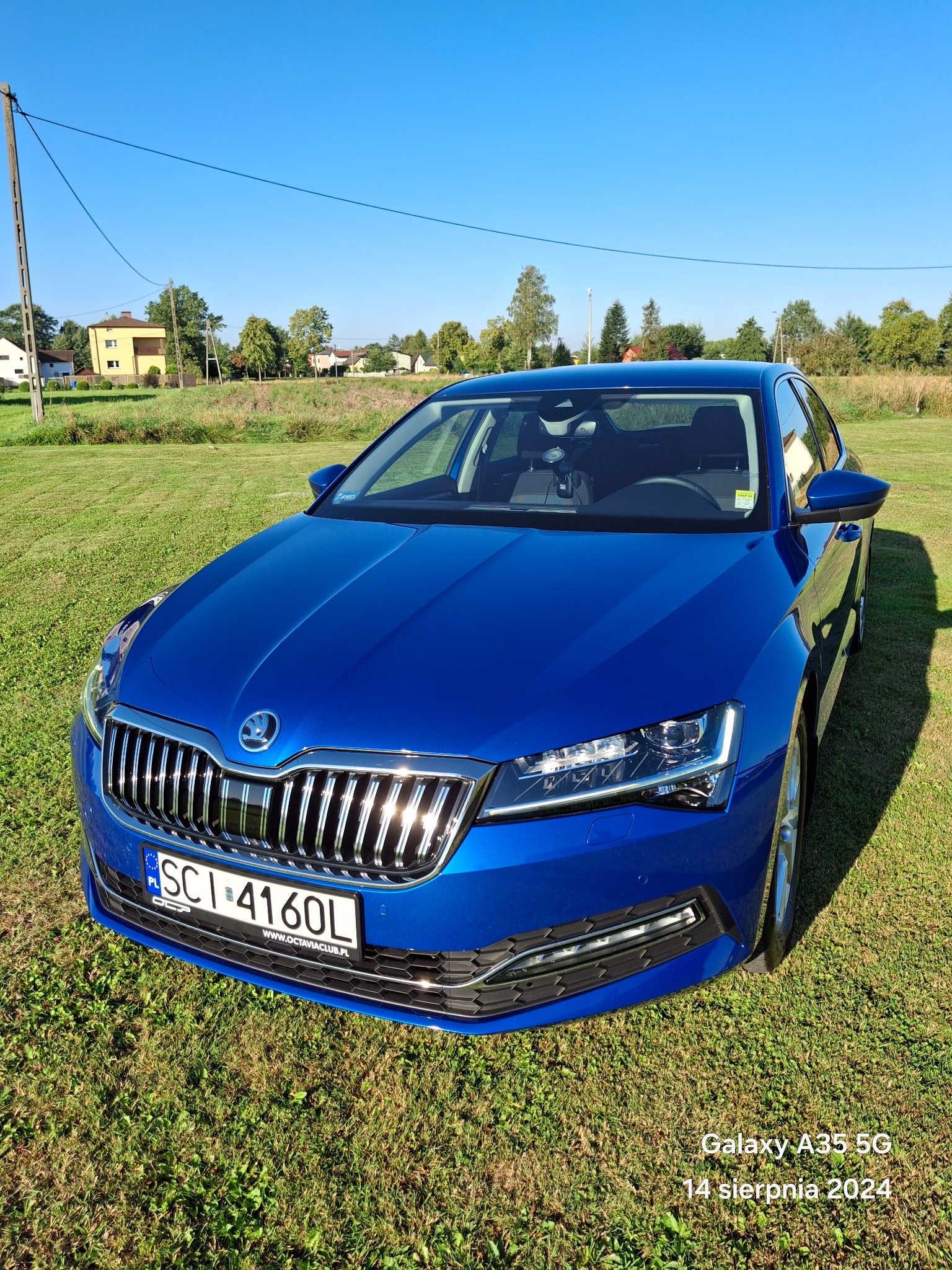 Niebieska Skoda Superb na trawniku w słoneczny dzień, widok z przodu, tablica rejestracyjna częściowo widoczna.
