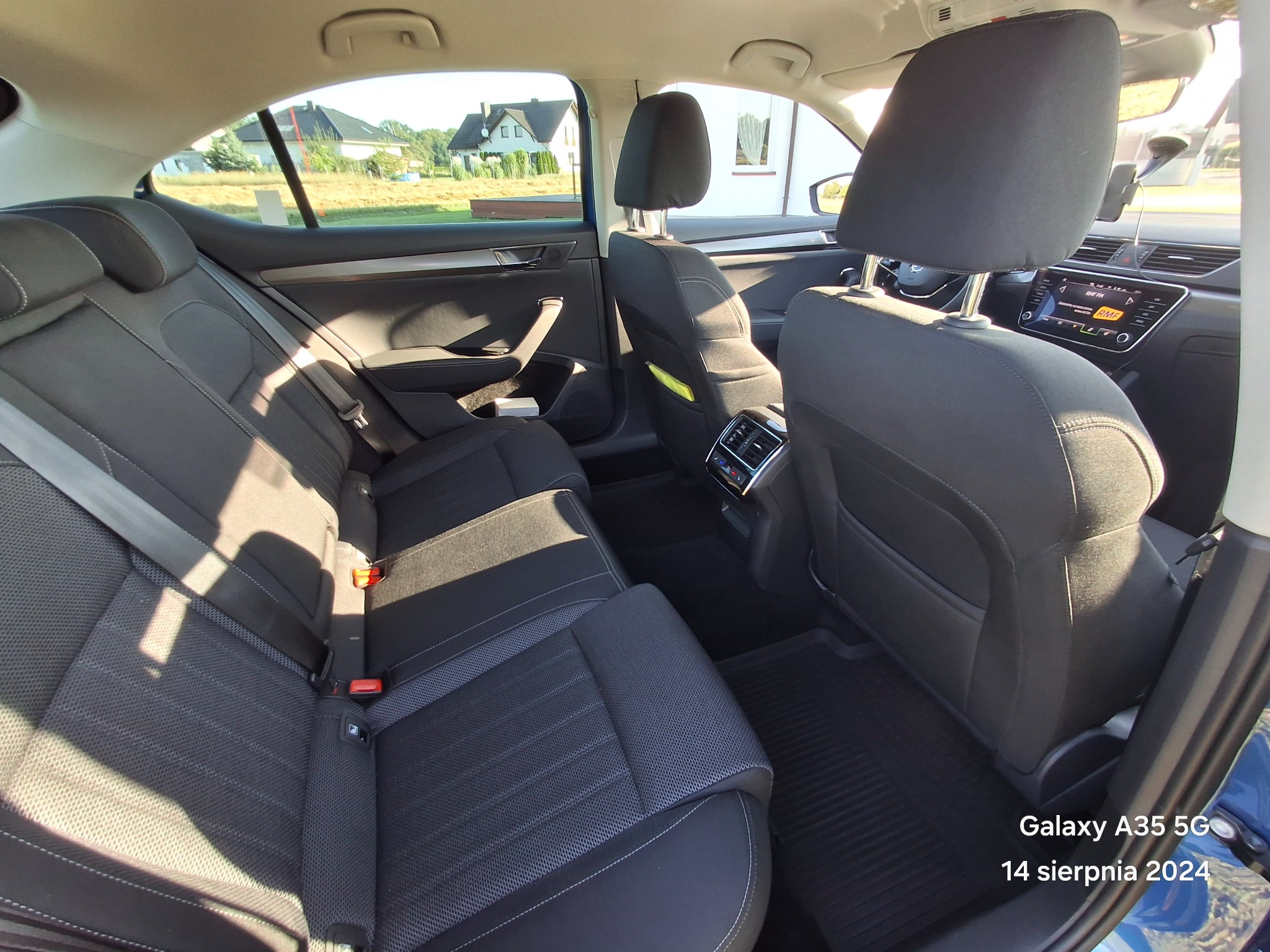 Wnętrze samochodu osobowego, widok na tylną kanapę z ciemną tapicerką, zagłówki, pasy bezpieczeństwa i kratki wentylacyjne, w tle widok na domy przez okno.