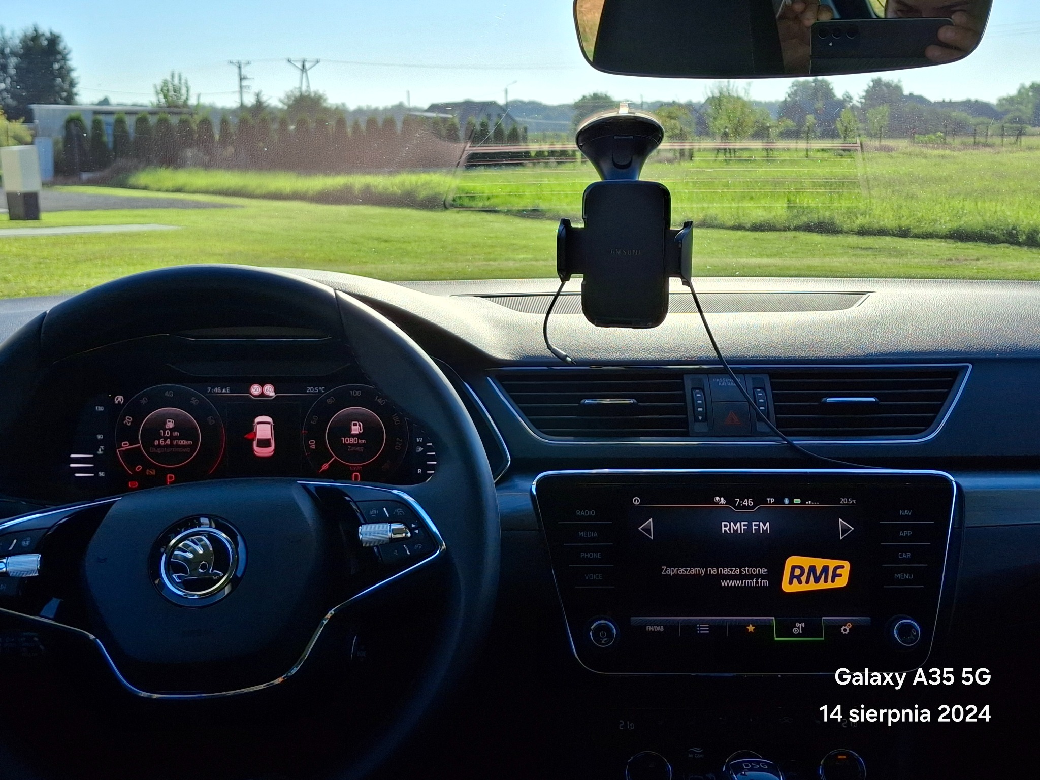 Wnętrze samochodu z widokiem na zielony krajobraz przez przednią szybę, uchwyt na telefon Samsung zamocowany na desce rozdzielczej, widoczne zegary i ekran radia RMF FM.