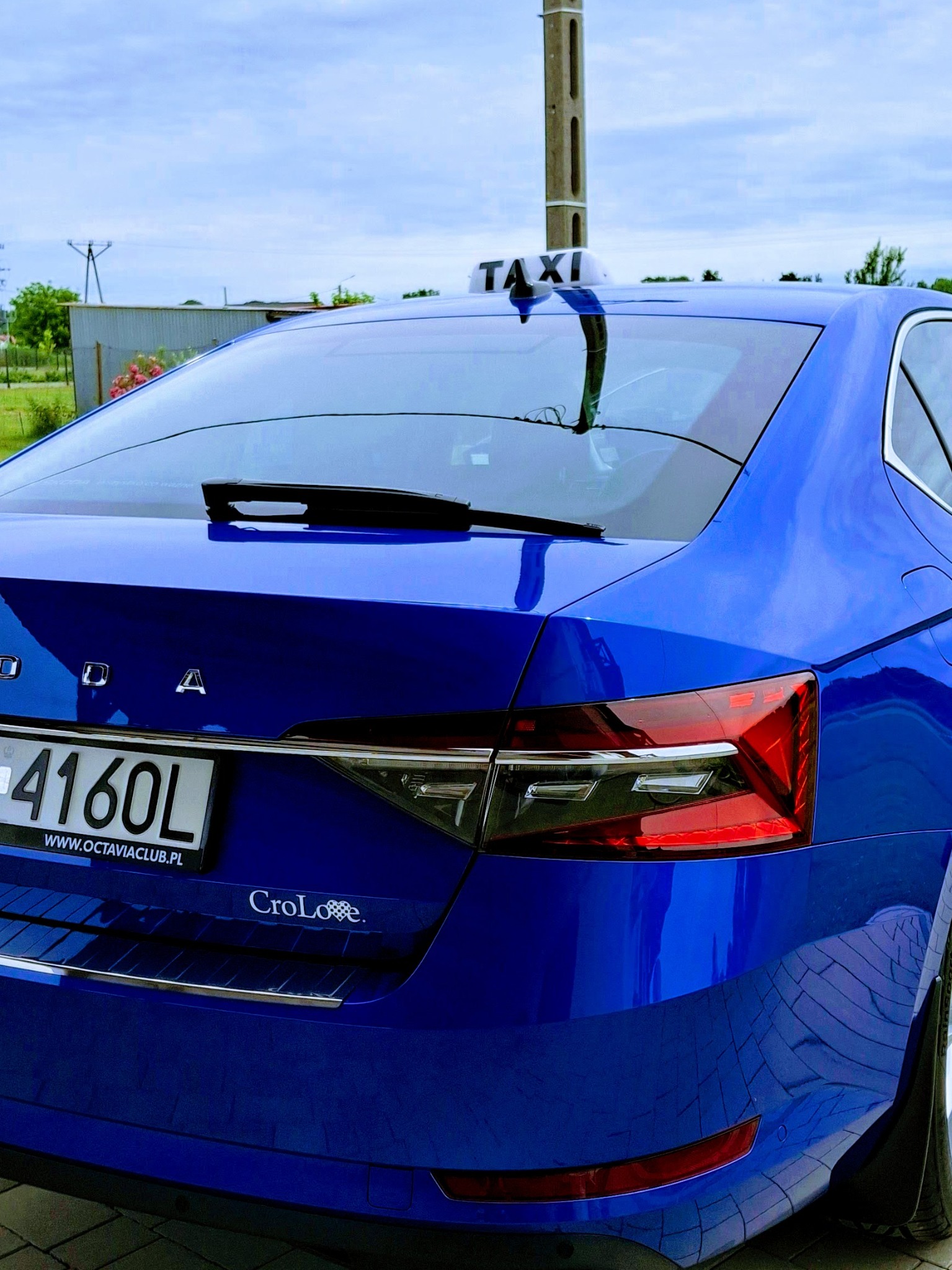 Niebieska Skoda Superb Taxi z tablicą rejestracyjną 4160L i napisem TAXI na dachu, widziana z tyłu na tle błękitnego nieba.
