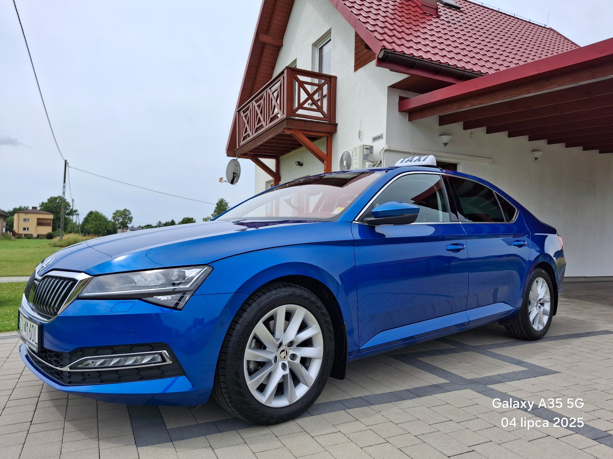 Niebieska taksówka Skoda Superb na tle domu z balkonem i czerwoną dachówką, zaparkowana na kostce brukowej. Widoczny napis 'TAXI' na dachu.