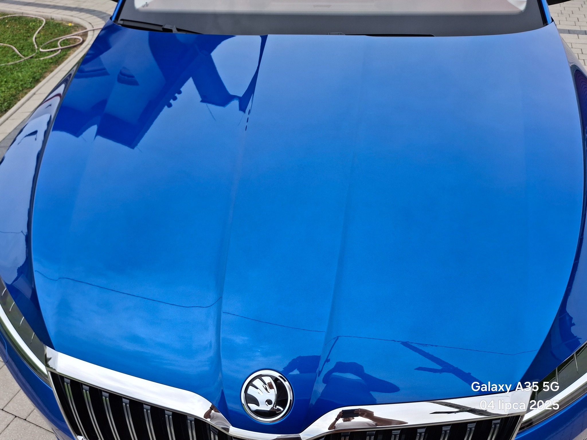 Błękitna maska samochodu marki Skoda, widok z góry, z połyskującym lakierem i chromowanym grillem. Widoczne odbicie osoby robiącej zdjęcie.