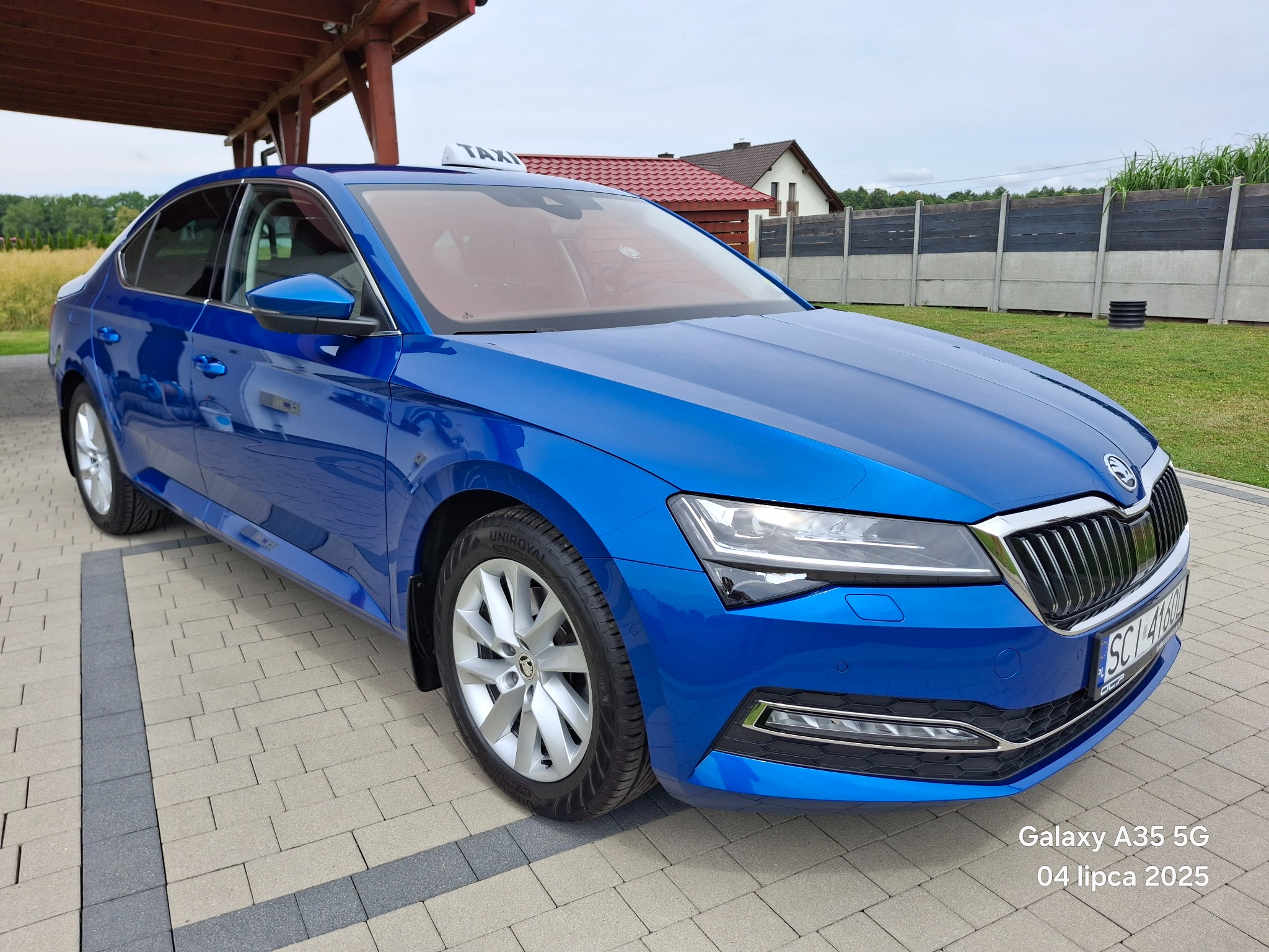 Błękitna Skoda Superb Taxi z napisem TAXI na dachu, zaparkowana na kostce brukowej, z widocznym fragmentem budynku w tle i zielenią.