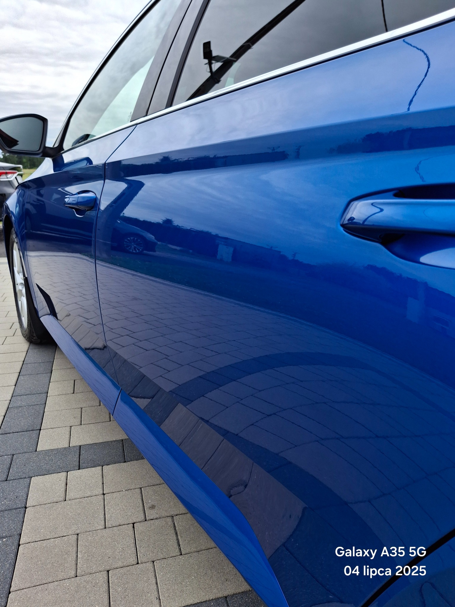 Błyszczący, błękitny lakier samochodu odbija chmury i kostkę brukową. Widoczny fragment drzwi, klamka i lusterko. Perspektywa z dołu podkreśla głębię koloru.
