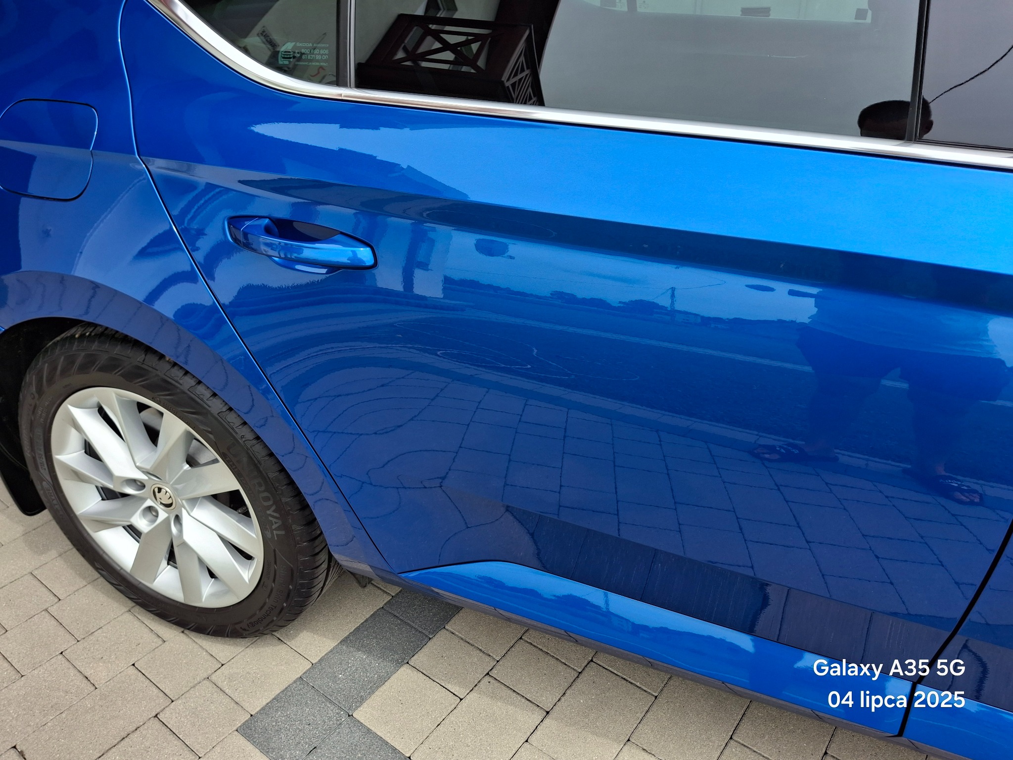 Błyszczący, niebieski samochód Skoda, widok z boku, z refleksami otoczenia na lakierze. Widoczne koło i fragment chodnika z kostki brukowej.