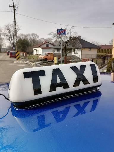 Lampa TAXI na niebieskim dachu samochodu w Katowicach, w tle znak drogowy TAXI i zabudowa jednorodzinna.