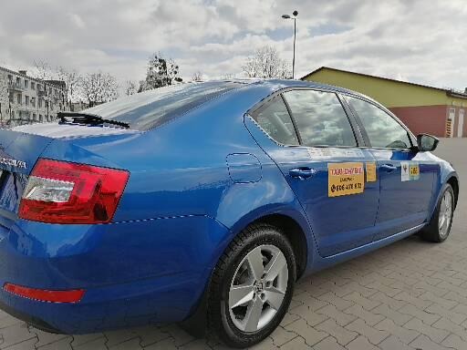 Niebieska taksówka Skoda Octavia z żółtymi naklejkami informacyjnymi na drzwiach, zaparkowana na szarym chodniku w pochmurny dzień.