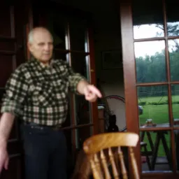 Mężczyzna w koszuli w kratę wskazuje palcem na zewnątrz przez otwarte, drewniane drzwi balkonowe z widokiem na zielony trawnik i drzewa.