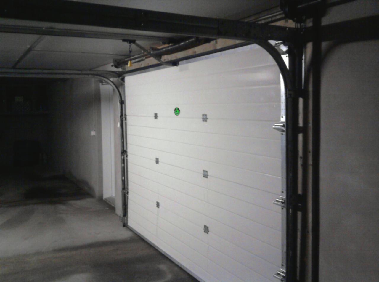 Biała segmentowa brama garażowa, widok od wewnątrz garażu. Metalowa konstrukcja prowadnic, poziome panele bramy z delikatnymi przetłoczeniami.