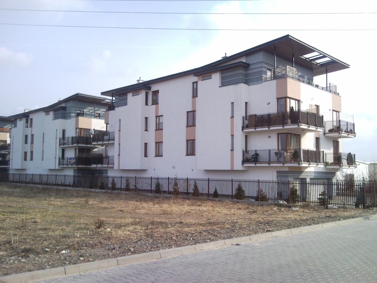 Nowoczesny budynek wielorodzinny z balkonami i metalową balustradą, widoczny z poziomu ulicy w słoneczny dzień. Minimalistyczna architektura.