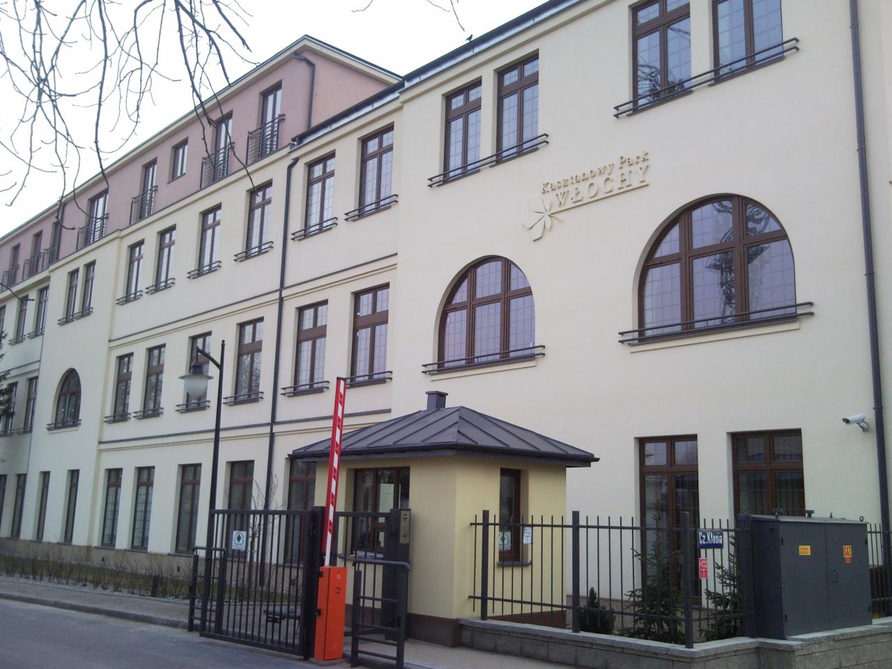 Budynek z bramą wjazdową i szlabanem, widoczne okna z brązowymi ramami i tabliczka z napisem 'Kaszatanowy Park Włochy'.
