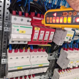 Sprawdzanie napięcia w rozdzielnicy elektrycznej za pomocą testera napięcia, widoczne bezpieczniki i przewody podłączone do szyny DIN.