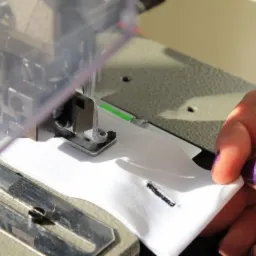 Detal maszyny do szycia dziurek w białej tkaninie, dłoń z fioletowym lakierem przytrzymuje materiał.