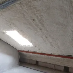 Poddasze po ociepleniu pianką, widoczne krokwie, okno dachowe, pianka o nierównej strukturze pokrywa całą powierzchnię, ściany boczne pomalowane na szaro.