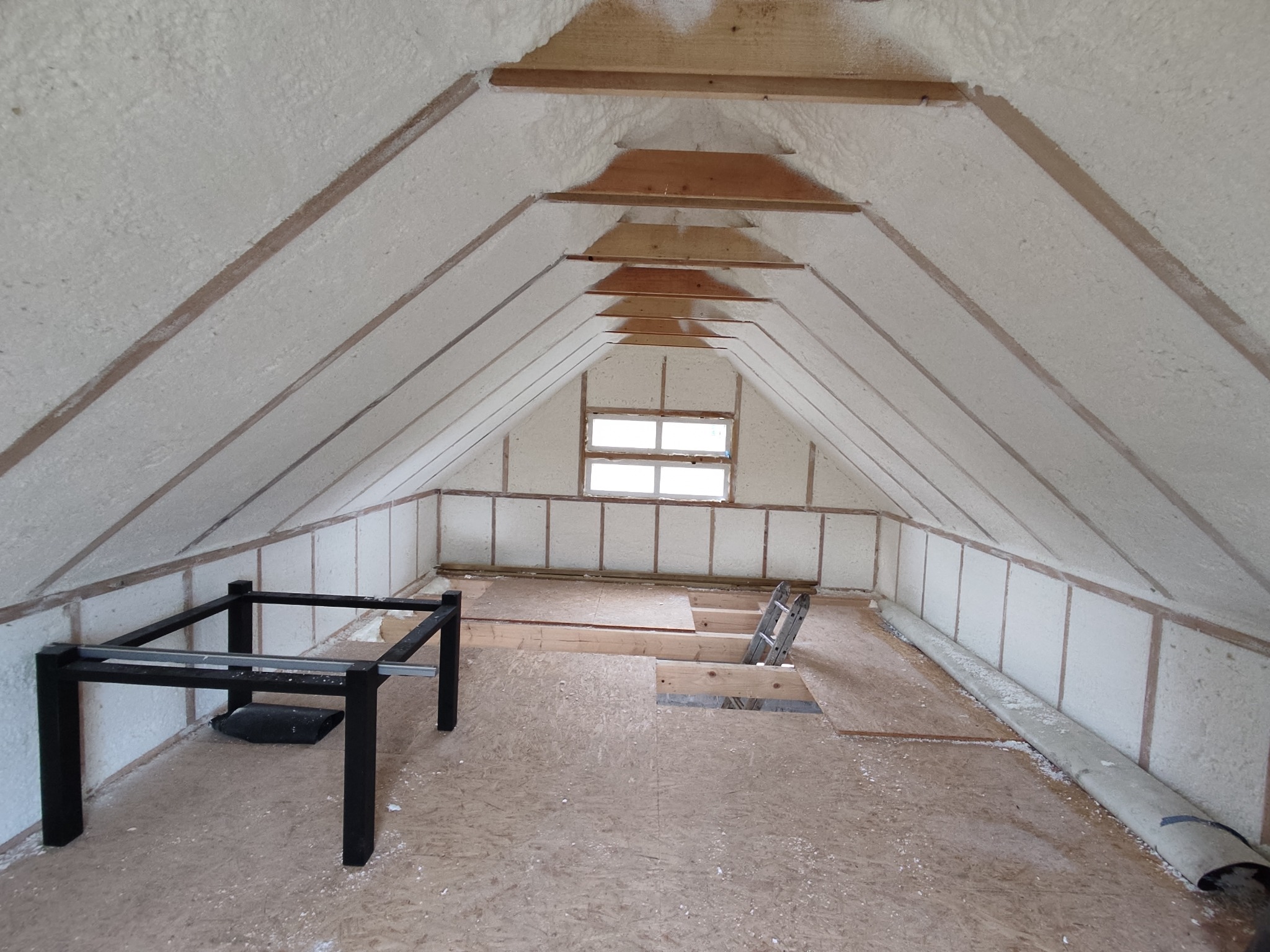 Poddasze z drewnianą konstrukcją pokryte białą pianką izolacyjną, widoczny fragment podłogi z płyty OSB i czarna, metalowa konstrukcja stołu.