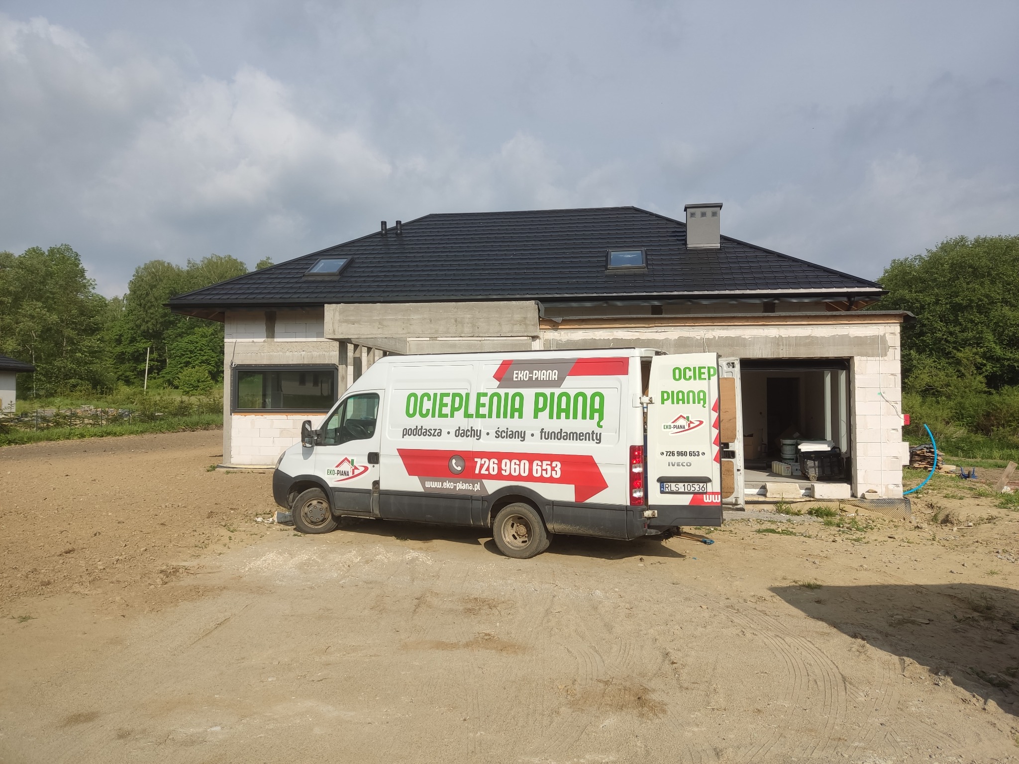 Biała furgonetka firmy EKO-PIANA z reklamą ocieplania pianą poddaszy, dachów, ścian i fundamentów, zaparkowana przed budynkiem w trakcie budowy z widocznym dachem i oknami dachowymi.