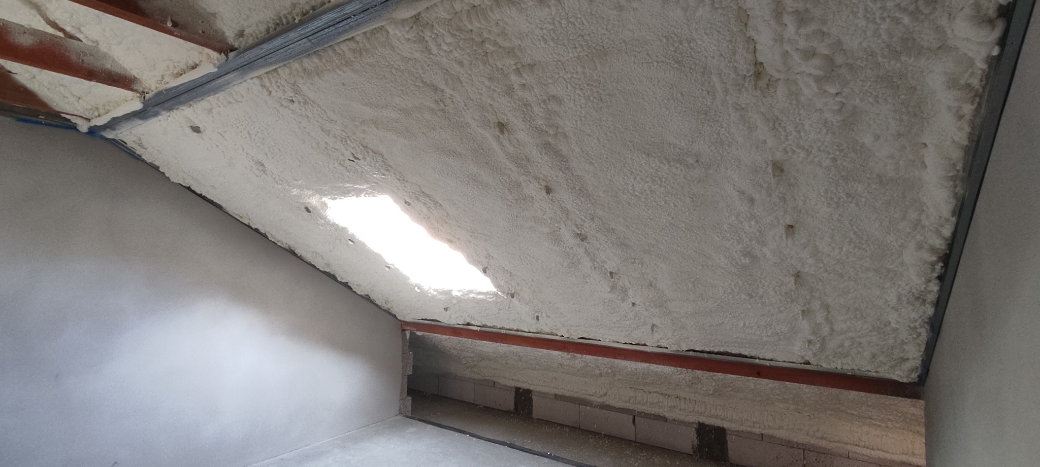 Poddasze po ociepleniu pianką, widoczne krokwie, okno dachowe, pianka o nierównej strukturze pokrywa całą powierzchnię, ściany boczne pomalowane na szaro.