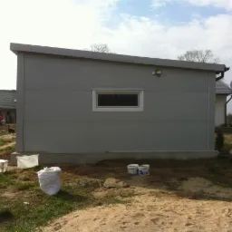 Szara hala z oknem na fundamencie, otoczona trawą i piaskiem. W tle dom z czerwonym autem. Widoczne puszki farby i worki. Hala z płyty warstwowej, lekki kąt dachu.