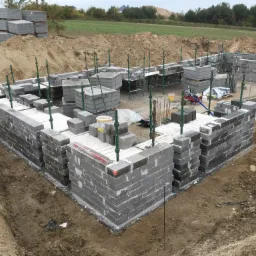 Wznoszenie ścian fundamentowych z bloczków betonowych na placu budowy, widoczne podpory szalunkowe i palety bloczków w tle.
