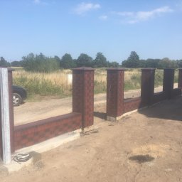 USŁUGI OGÓLNOBUDOWLANE ED-BUD EDWIN SZEFKA - Częściowo wzniesione ogrodzenie z cegły klinkierowej z metalowym słupkiem, widoczne przewody elektryczne, w tle pole i drzewa.